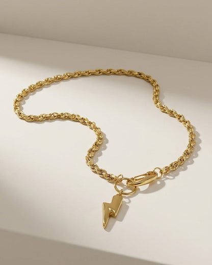 A gold-tone twisted rope chain necklace with a small lightning bolt pendant on a plain surface.
