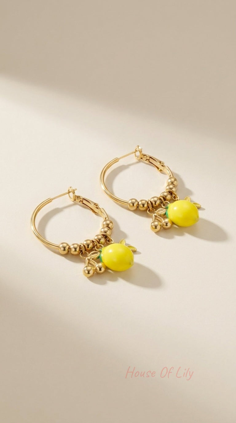 Gold hoop earrings featuring beaded detailing and dangling lemon charms on a neutral beige background.