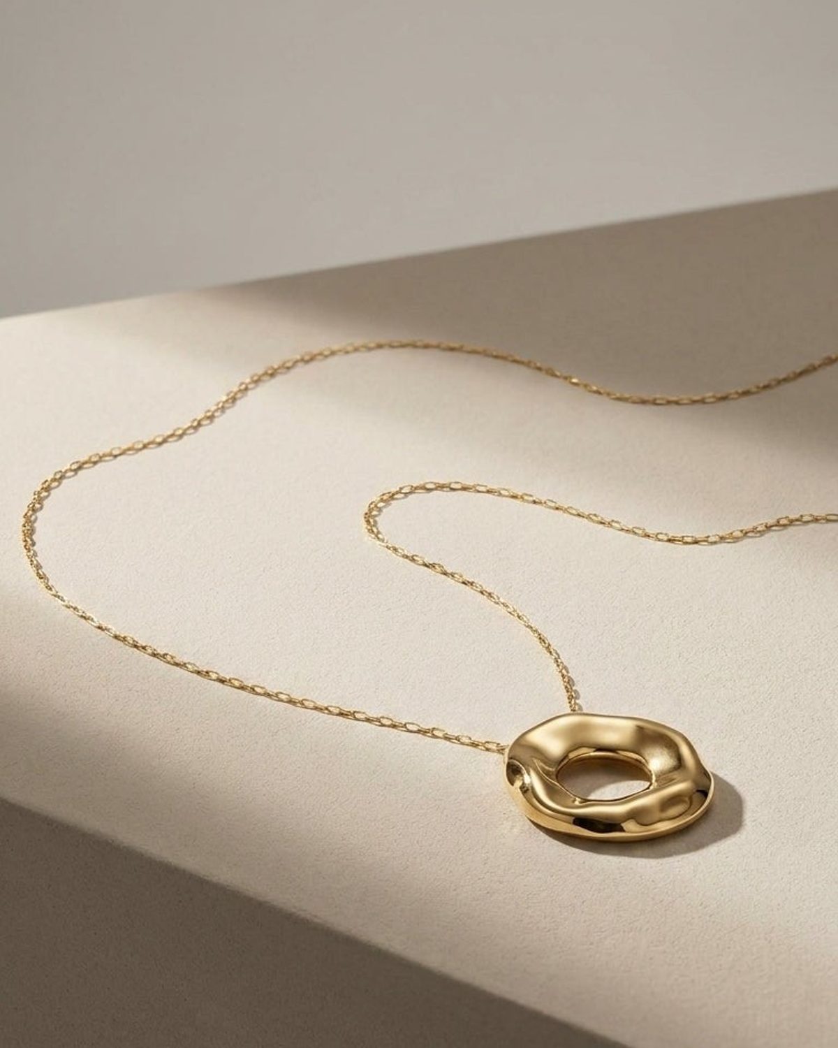 A gold-toned chain necklace with an irregular, organic-shaped circular pendant rests on a neutral surface.