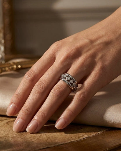 A silver ring with embedded rhinestones worn on a person's ring finger over a satin surface.