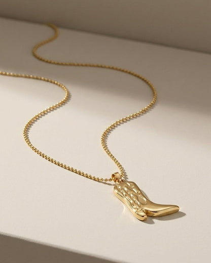 A gold cowboy boot pendant hangs from a matching beaded chain on a neutral surface.