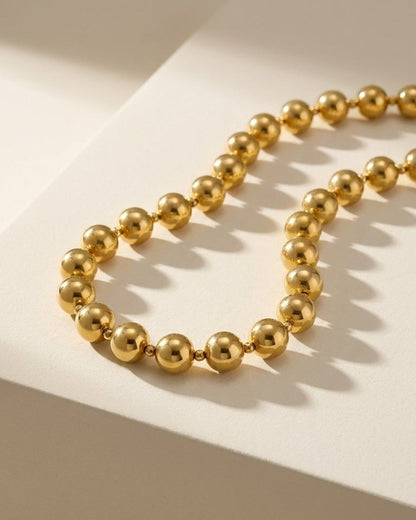 Gold beaded chain necklace rests on a neutral surface, casting soft shadows.