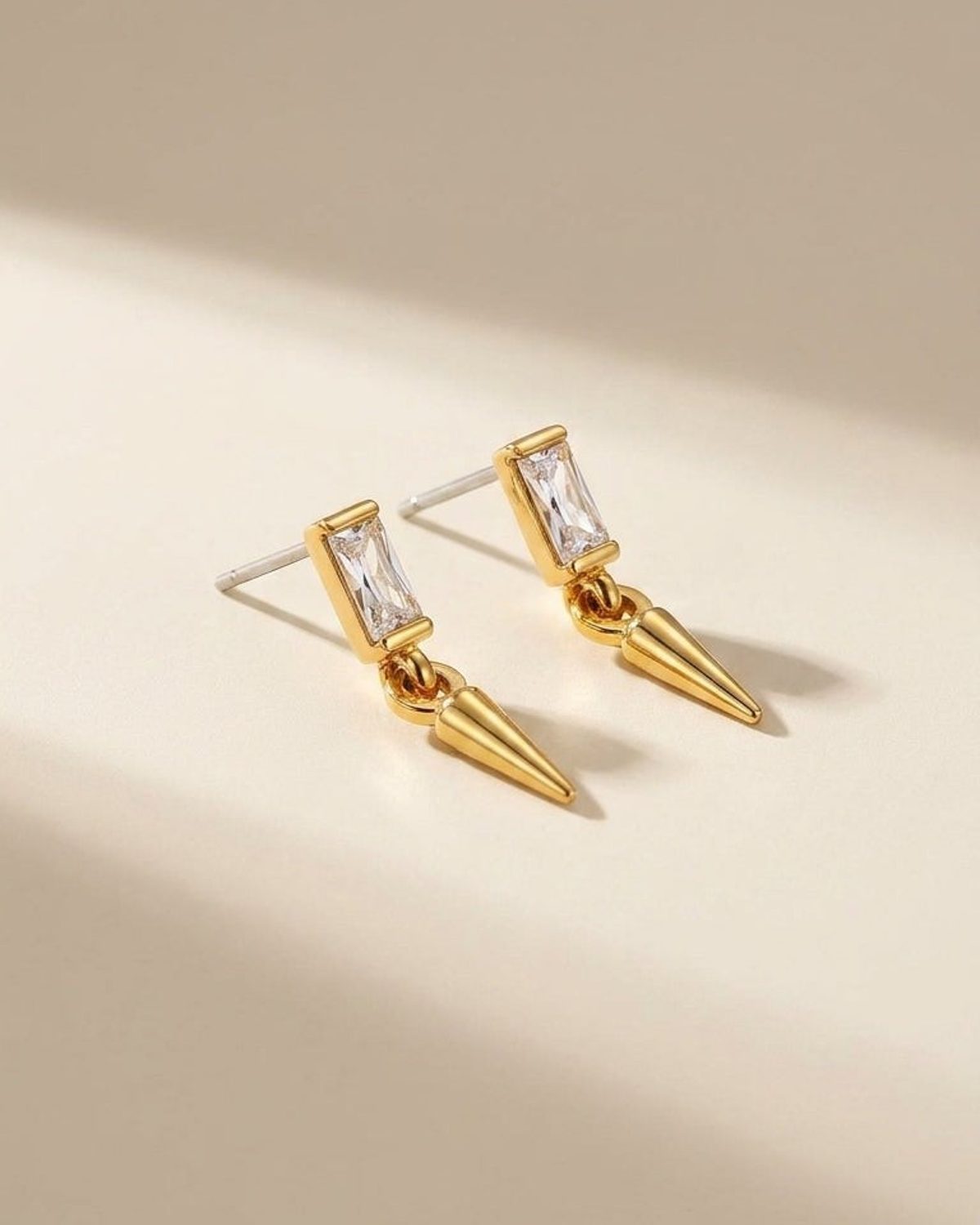 Gold-plated stud earrings with a rectangular clear crystal and a dangling cone charm on a neutral surface.