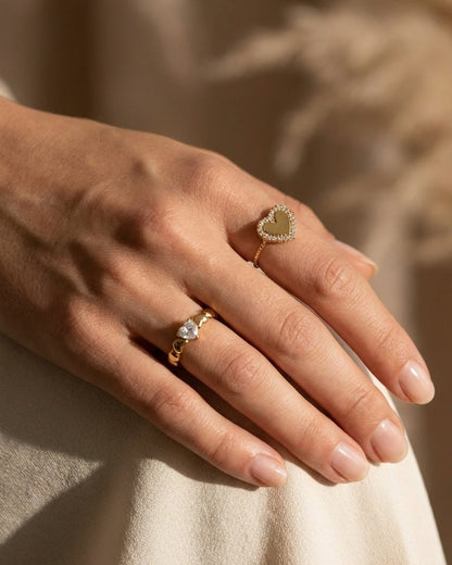 Gold rings on a hand: one features a zircon-studded heart frame, the other a heart-shaped stone.