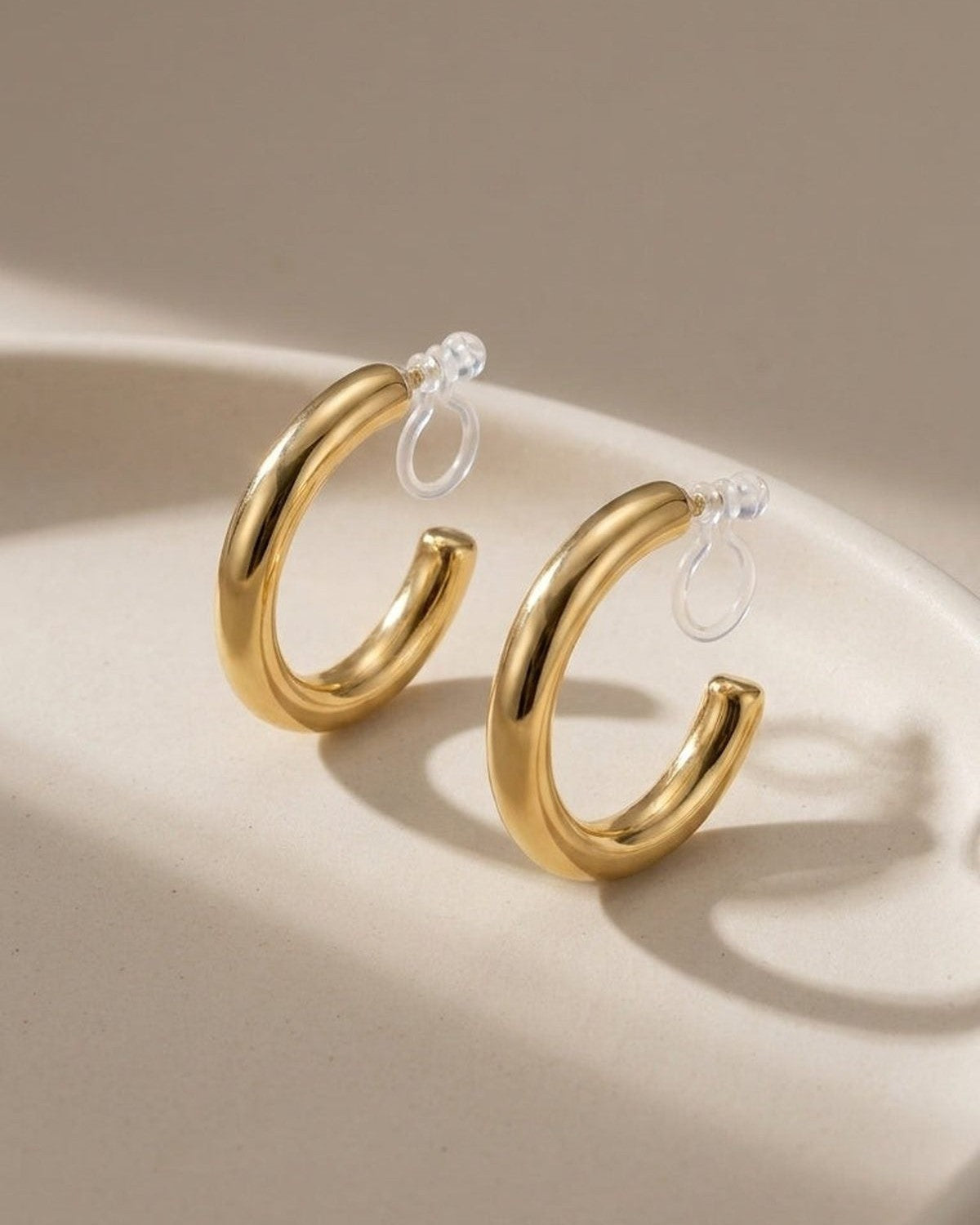 Thick gold hoop earrings with clear clip-on backs on a beige tray.