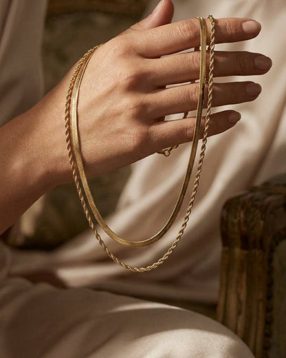 A hand holds a dual gold chain necklace with a flat herringbone chain and a twisted rope chain.