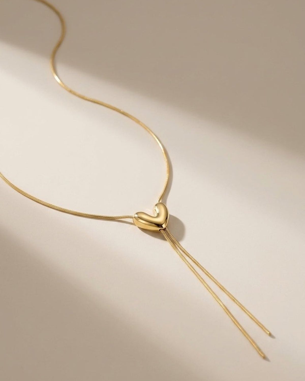 Gold-colored steel lariat necklace featuring a puffed heart pendant with two hanging chain strands.