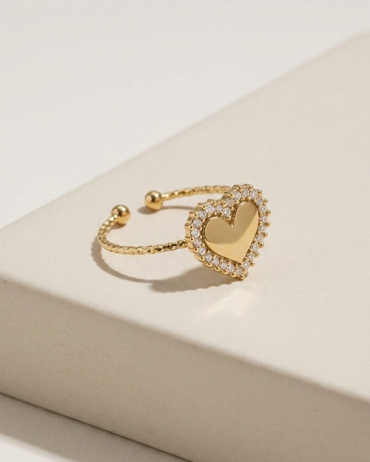 Gold adjustable ring with a heart-shaped centerpiece bordered by small clear stones, resting on a neutral surface.