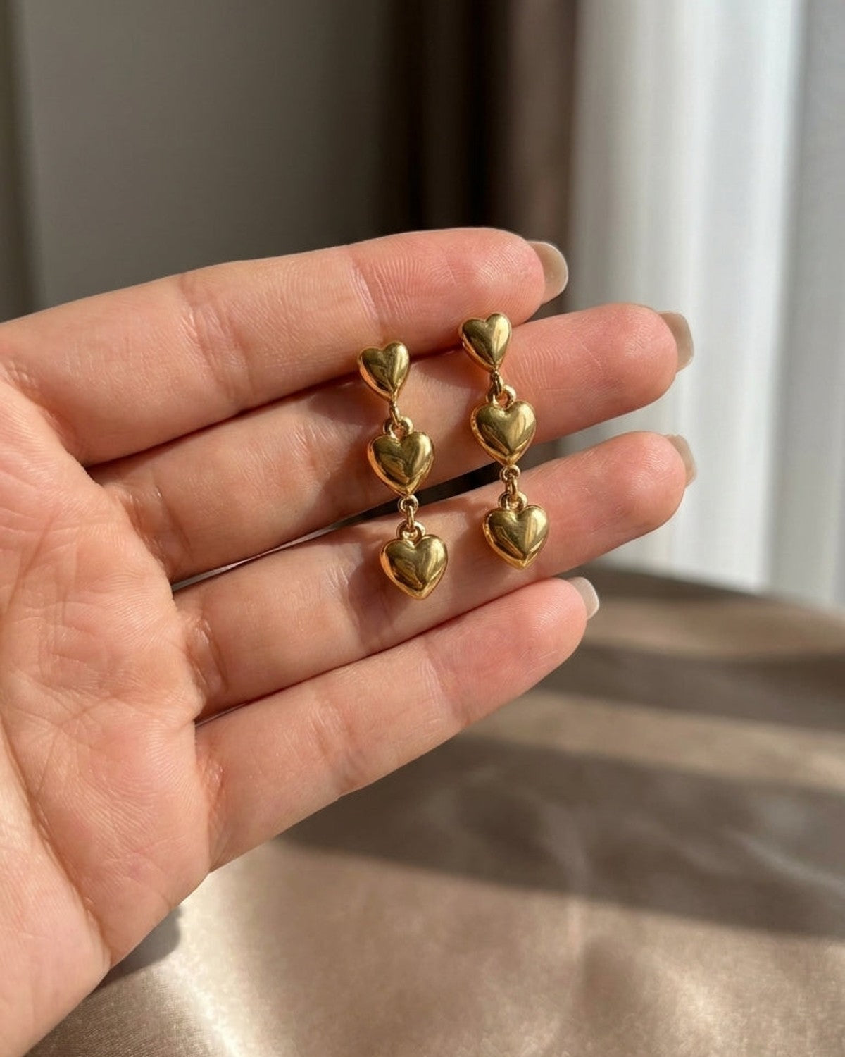 Two gold Heart Dangle Earrings resting on an open palm, each featuring three graduated puffy hearts linked together.
