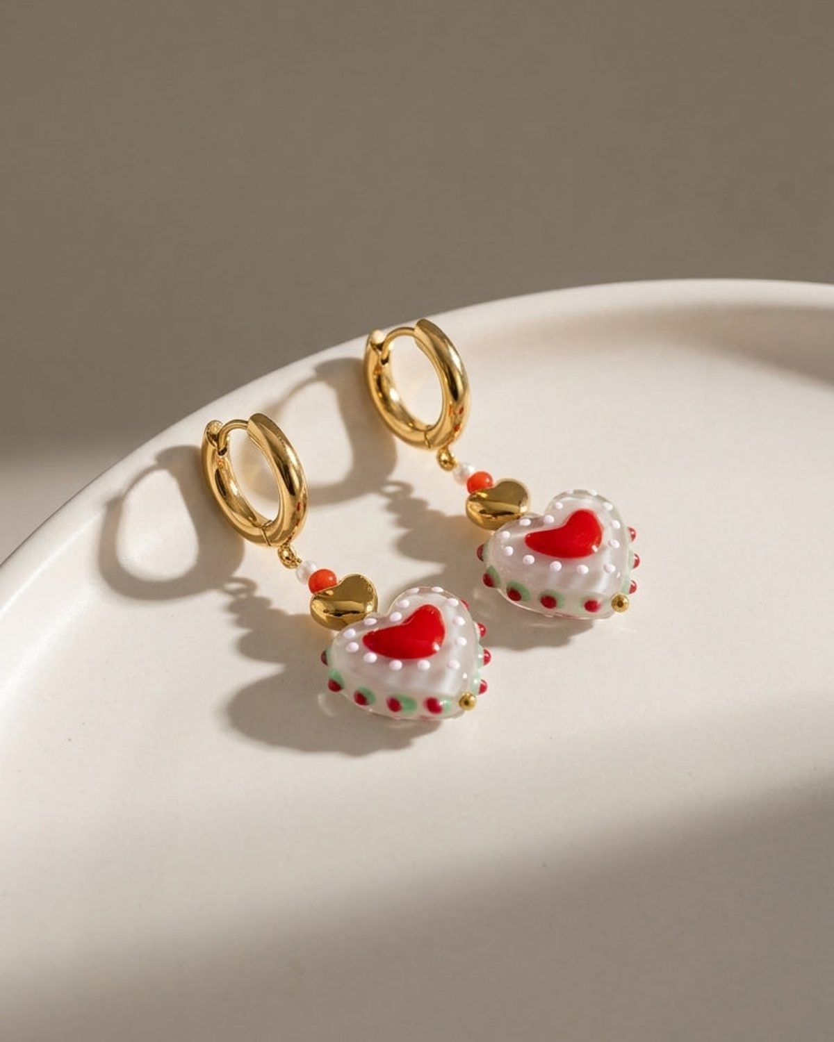 Gold hoop earrings with dangling white heart-shaped glass beads featuring red and green details.