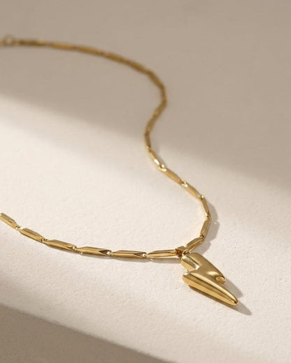 Gold lightning bolt pendant on a geometric-link chain, resting on a neutral surface.