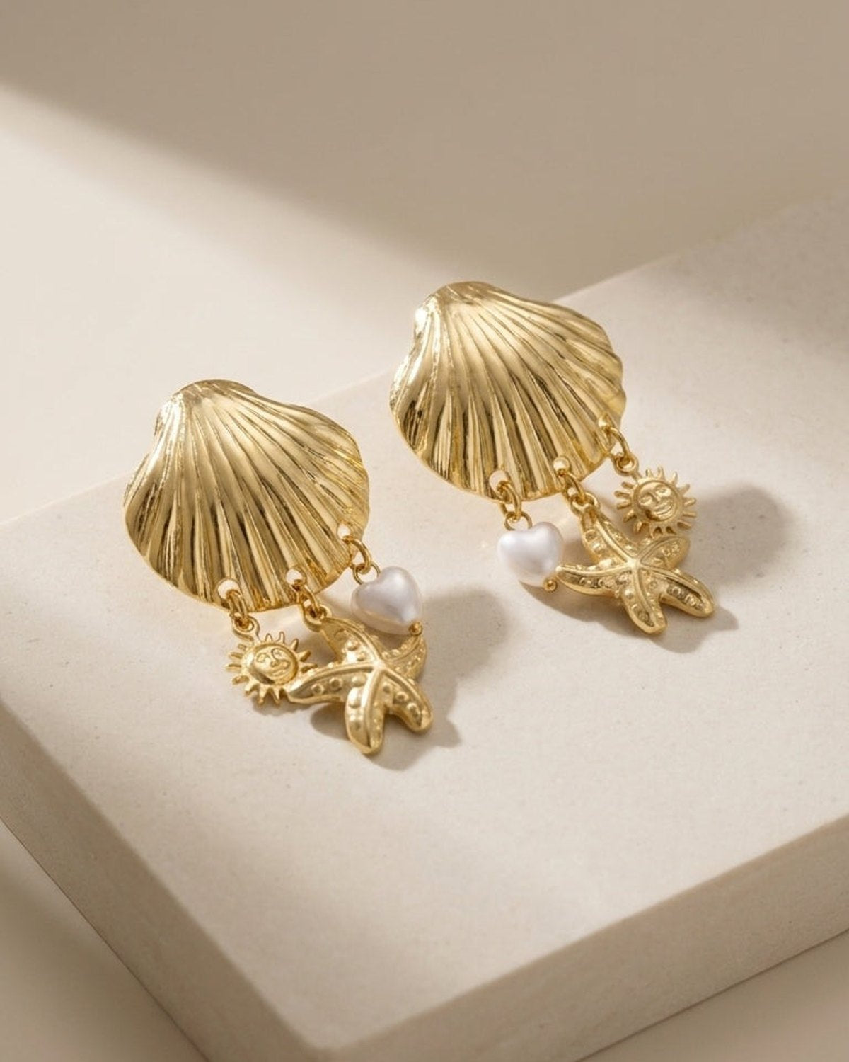 Gold-tone dangle earrings featuring a large shell, small sun, starfish, and a white heart charm on a stone base.