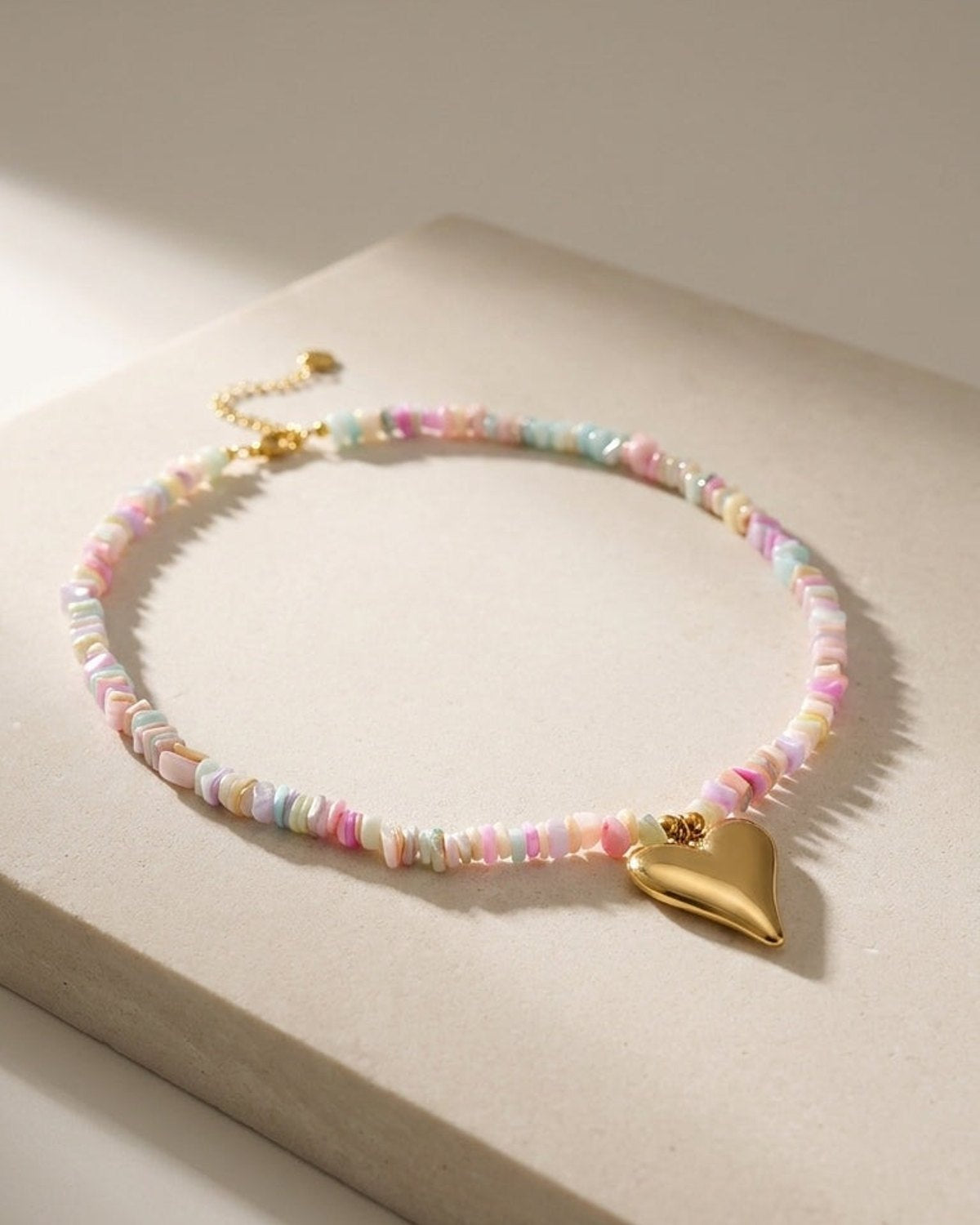 A necklace of multicolored pastel chipped beads with a smooth gold heart pendant on a neutral surface.