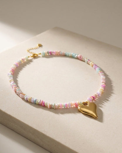A necklace of multicolored pastel chipped beads with a smooth gold heart pendant on a neutral surface.