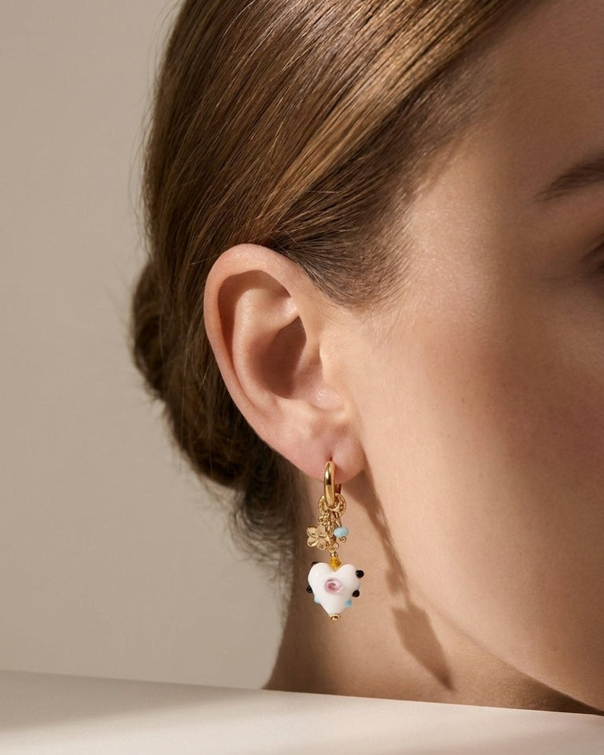 Gold hoop drop earring on an ear, featuring a white heart charm with pink and black details.