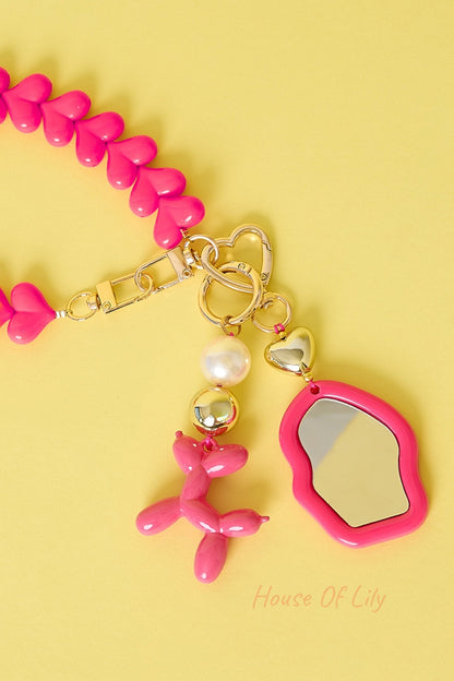 Pink heart-shaped chain bag charm with pink balloon dog, pearl and gold beads, and pink framed mirror keychain on yellow background