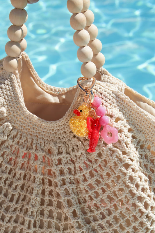 Beaded yellow duck keychain combined with pink and red charms hanging on a beige crocheted bag handle by a pool.