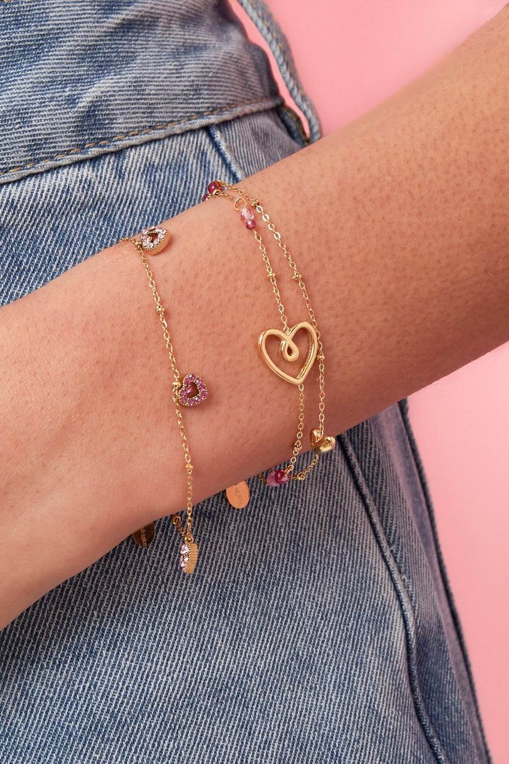 Gold chain bracelet on a wrist, featuring a hammered heart link and small pink pavé heart charms.
