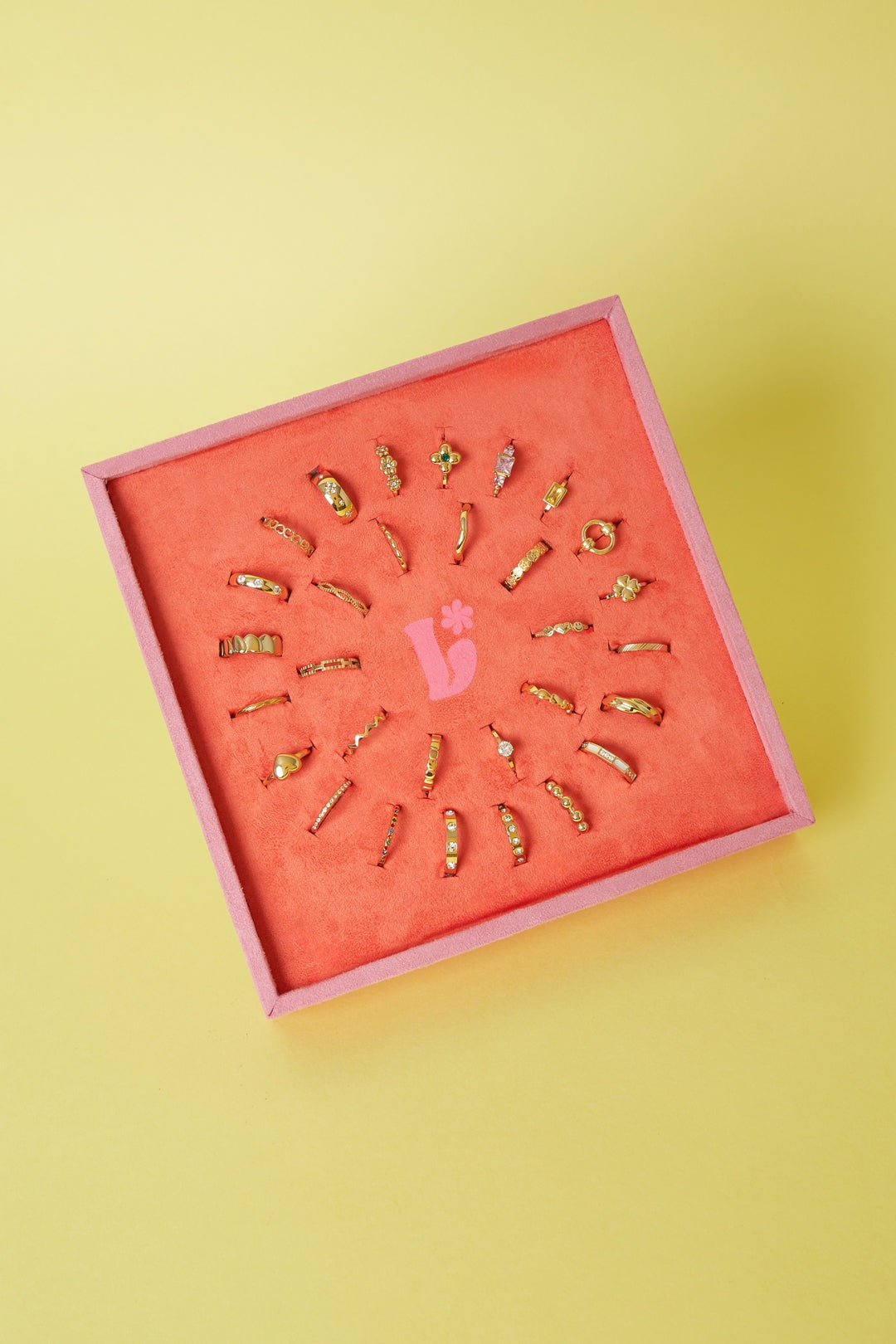 Angled view of coral-orange velvet display with 28 gold-tone rings showing various delicate designs including flowers and gemstones