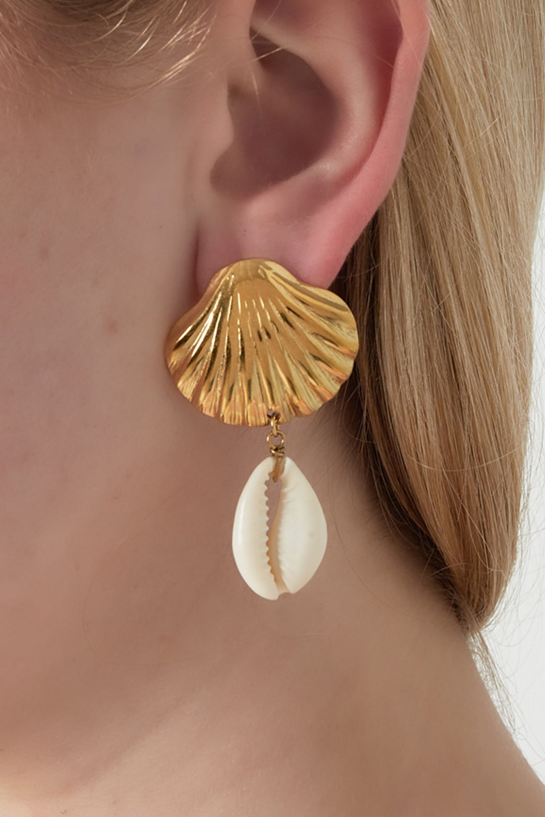 Close-up of gold seashell drop earring with white cowrie shell worn by a woman with light skin and blonde hair