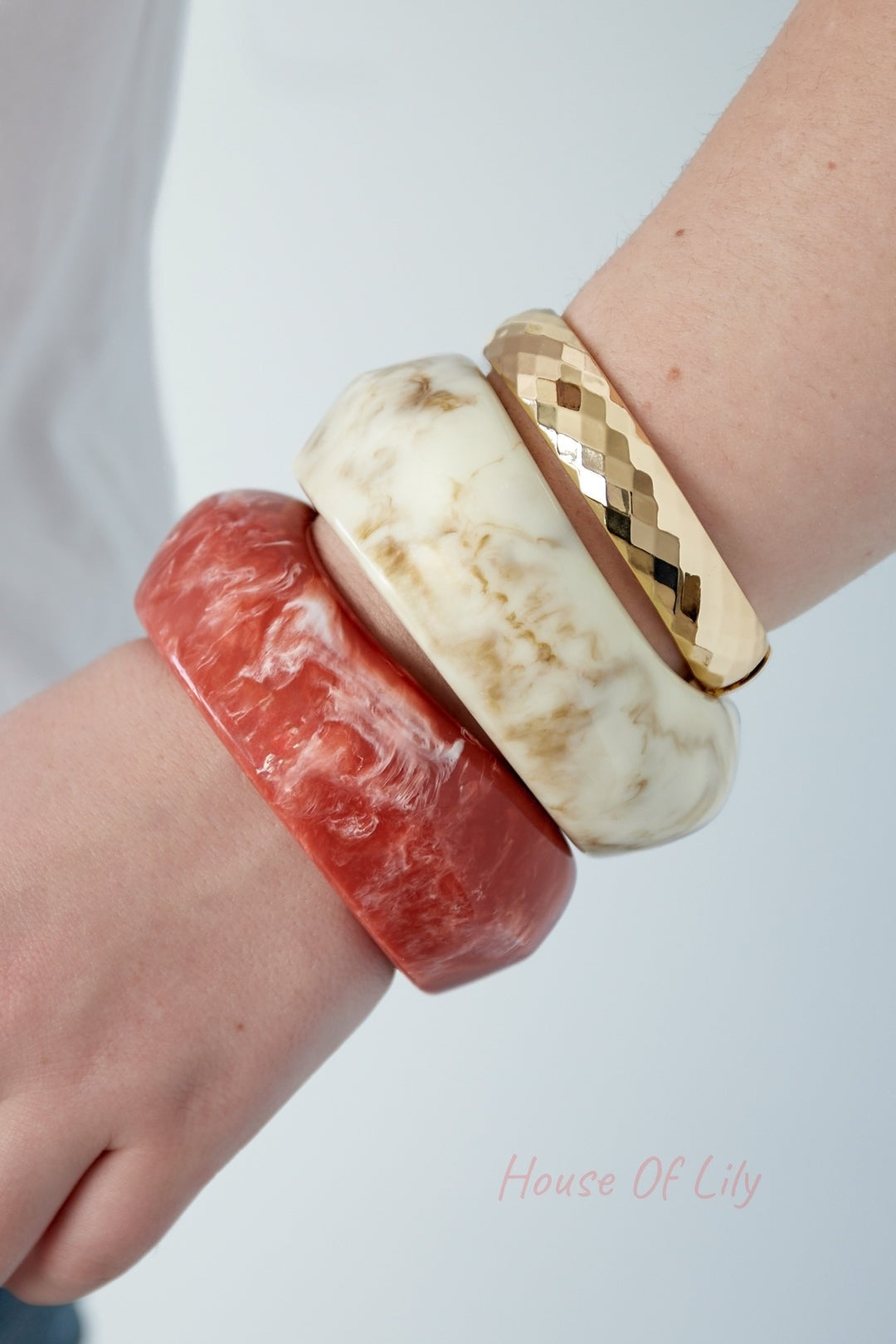 Human wrist wearing stacked bangles including marbled beige acrylic chunky bangle, coral marbled bangle, and gold metallic bangle
