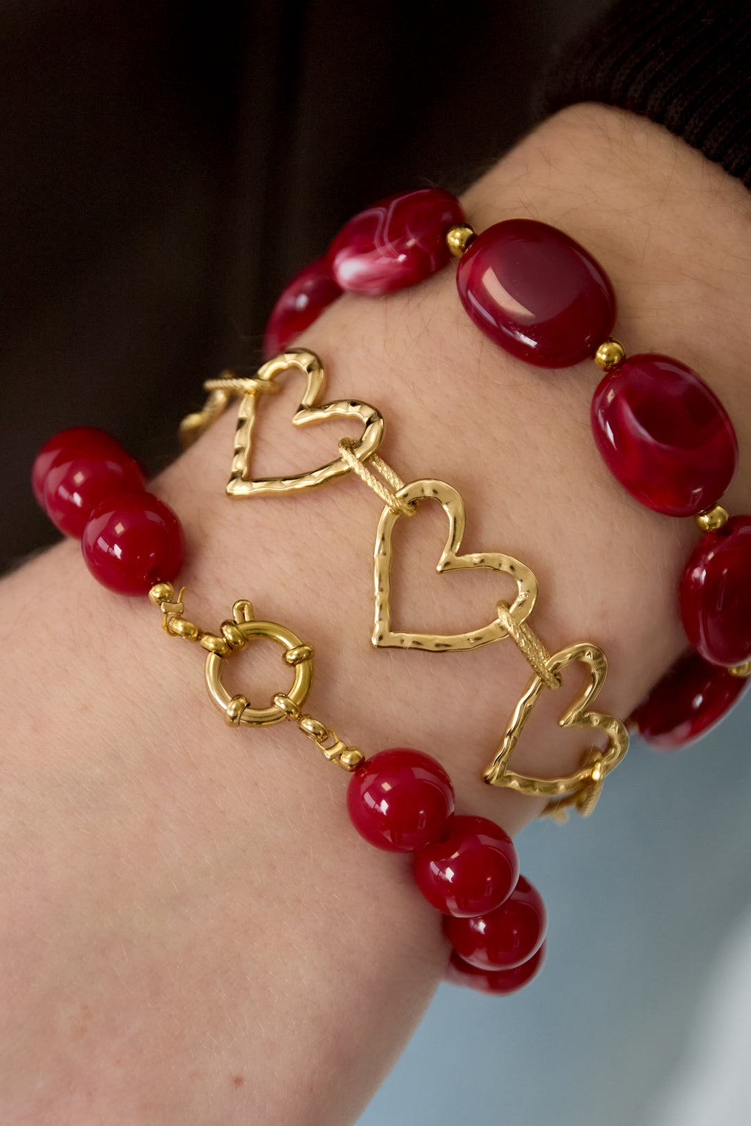 Wrist wearing two red beaded bracelets and one gold-tone heart link bracelet showing layered bracelet styling
