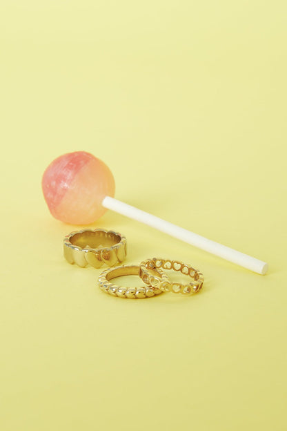 Three textured gold-tone rings arranged beside a pink and orange lollipop on a yellow background