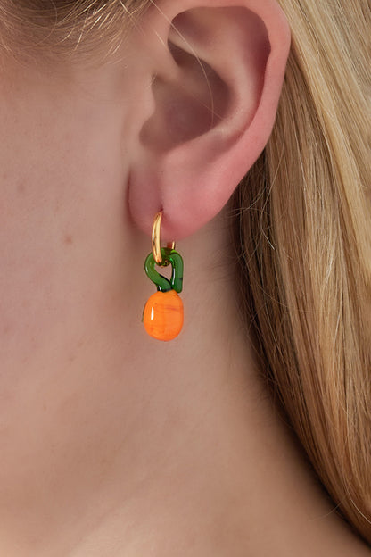 Close-up of a woman wearing a gold-plated hoop earring with a green loop and orange resin fruit charm, showcasing a playful drop earring style