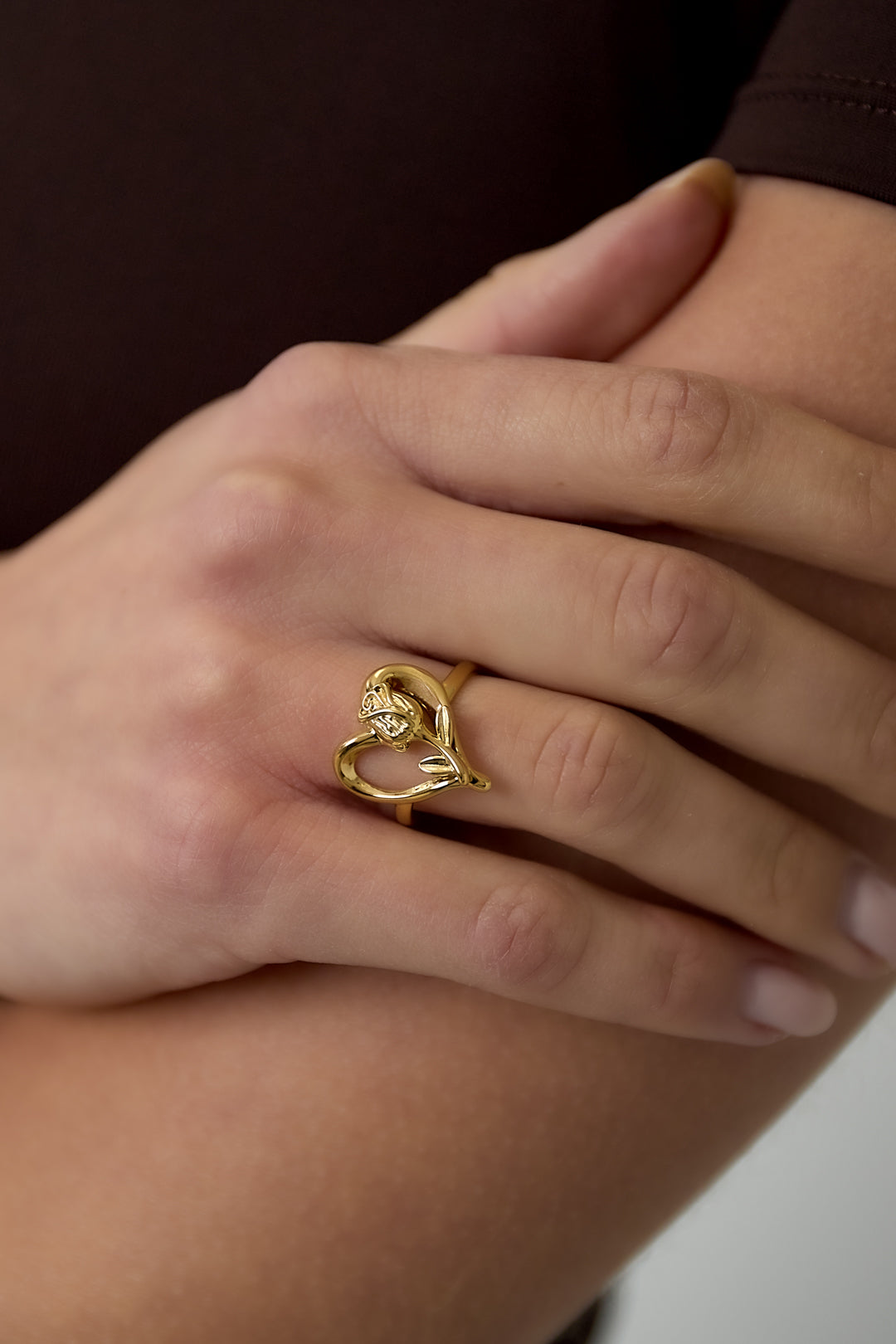 Close-up of hand wearing a gold rose heart ring with intertwined heart and rose motif, against soft skin tone