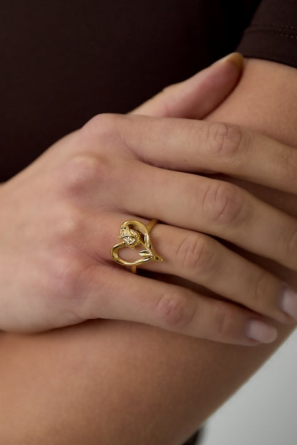 Close-up of hand wearing a gold rose heart ring with intertwined heart and rose motif, against soft skin tone