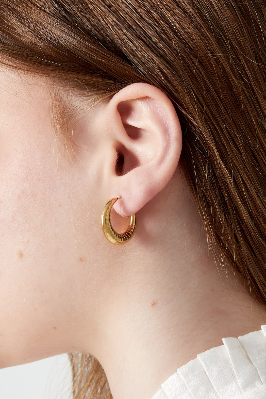 Close-up side view of a woman wearing one chunky gold clip-on hoop earring with ruffled white blouse