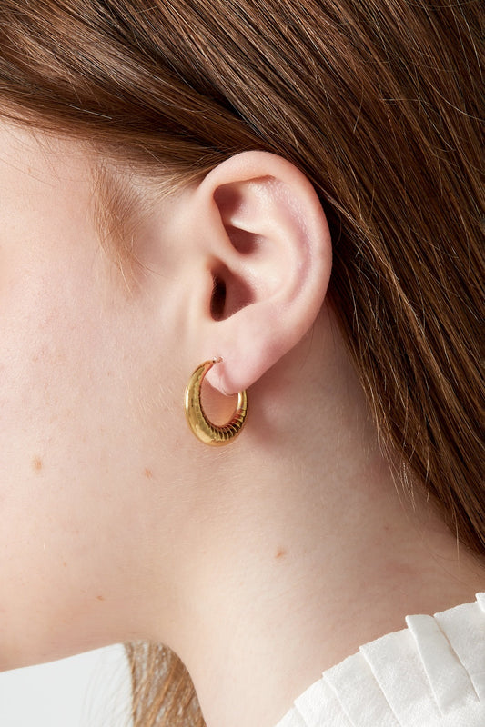 Close-up side view of a woman wearing one chunky gold clip-on hoop earring with ruffled white blouse
