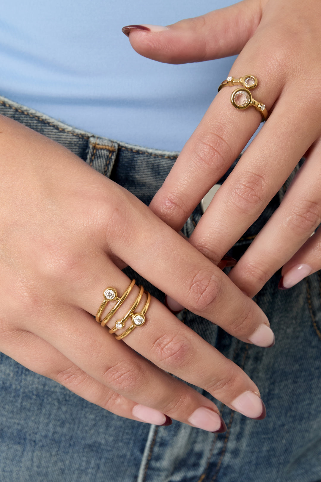 Close-up of model’s hand wearing a gold tone multi-band statement ring with rhinestones paired with casual denim and blue top