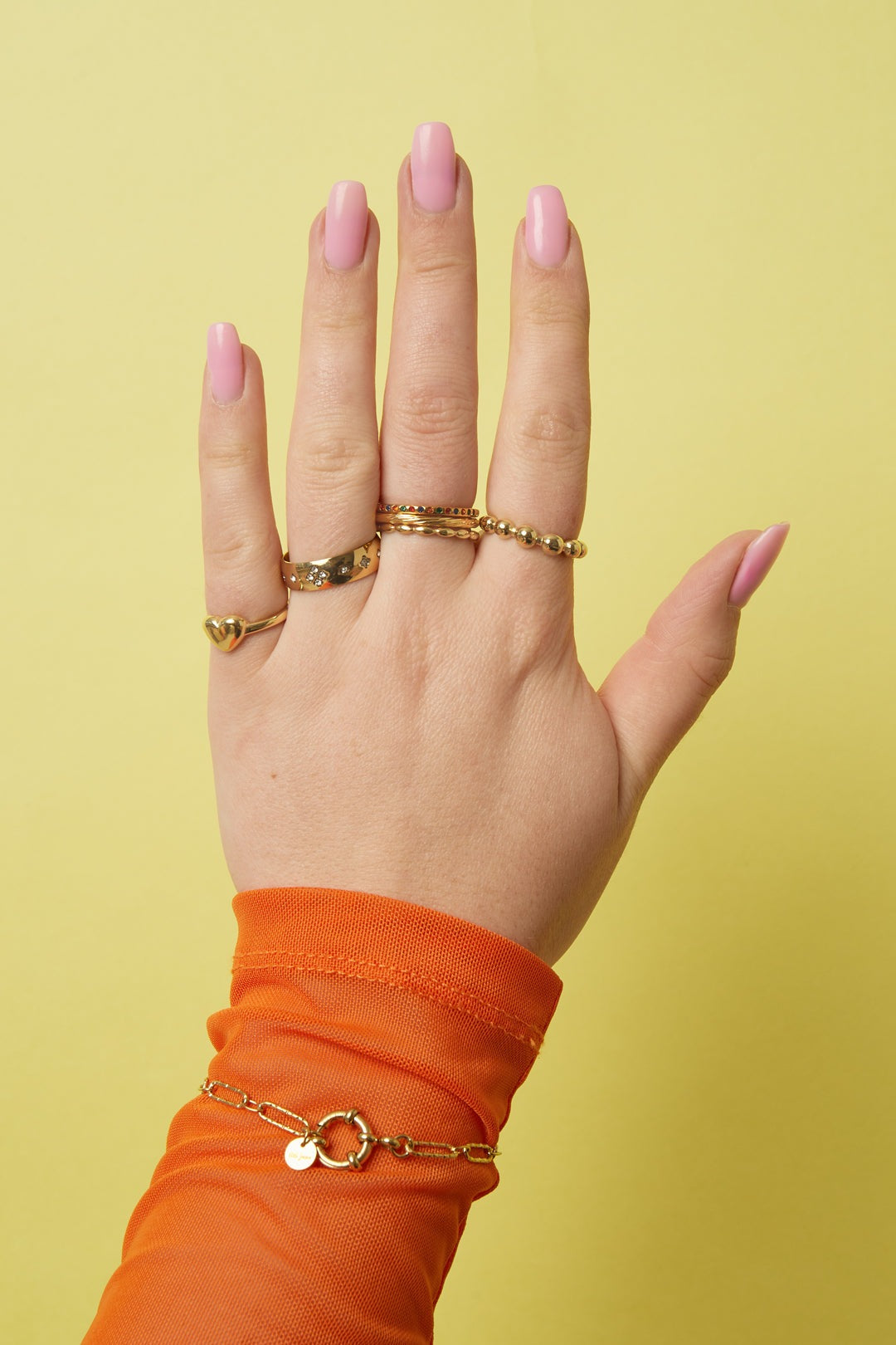 Hand wearing four gold rings including a beaded gold ring, displayed against a yellow background with pink nail polish and orange sleeve