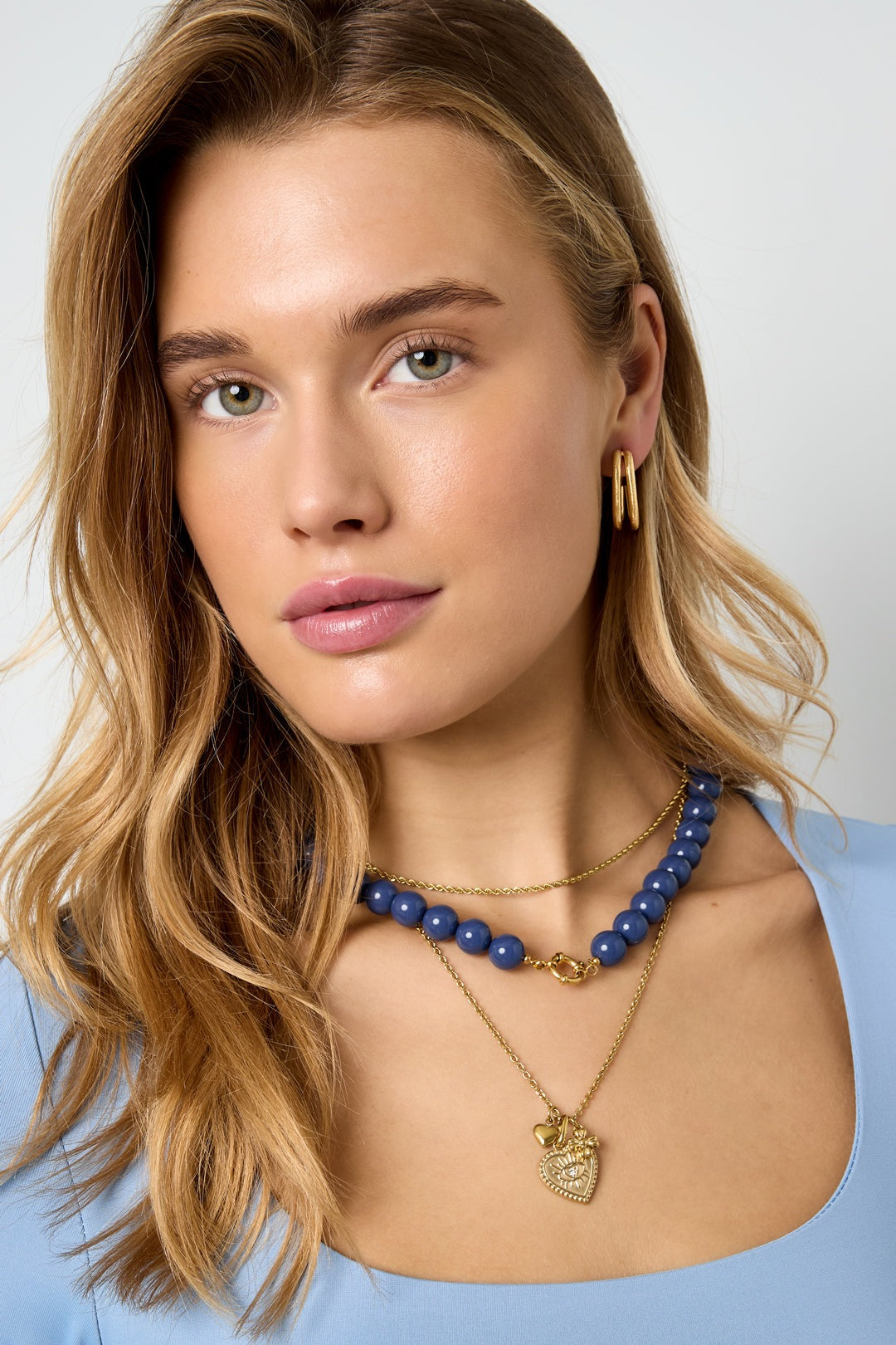 Model wearing layered necklaces including chunky navy blue beaded necklace with gold clasp and two gold chains paired with gold hoop earrings