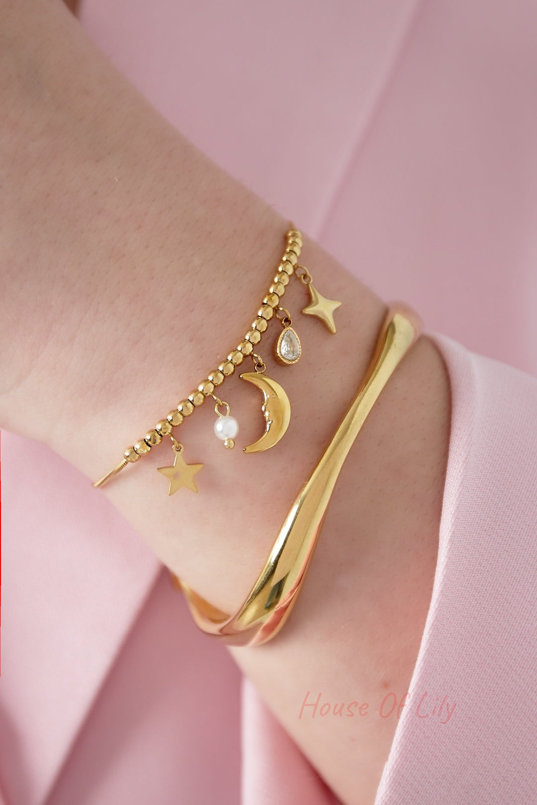 Close-up of gold celestial charm bracelet worn alongside a modern gold cuff on a wrist in pink clothing