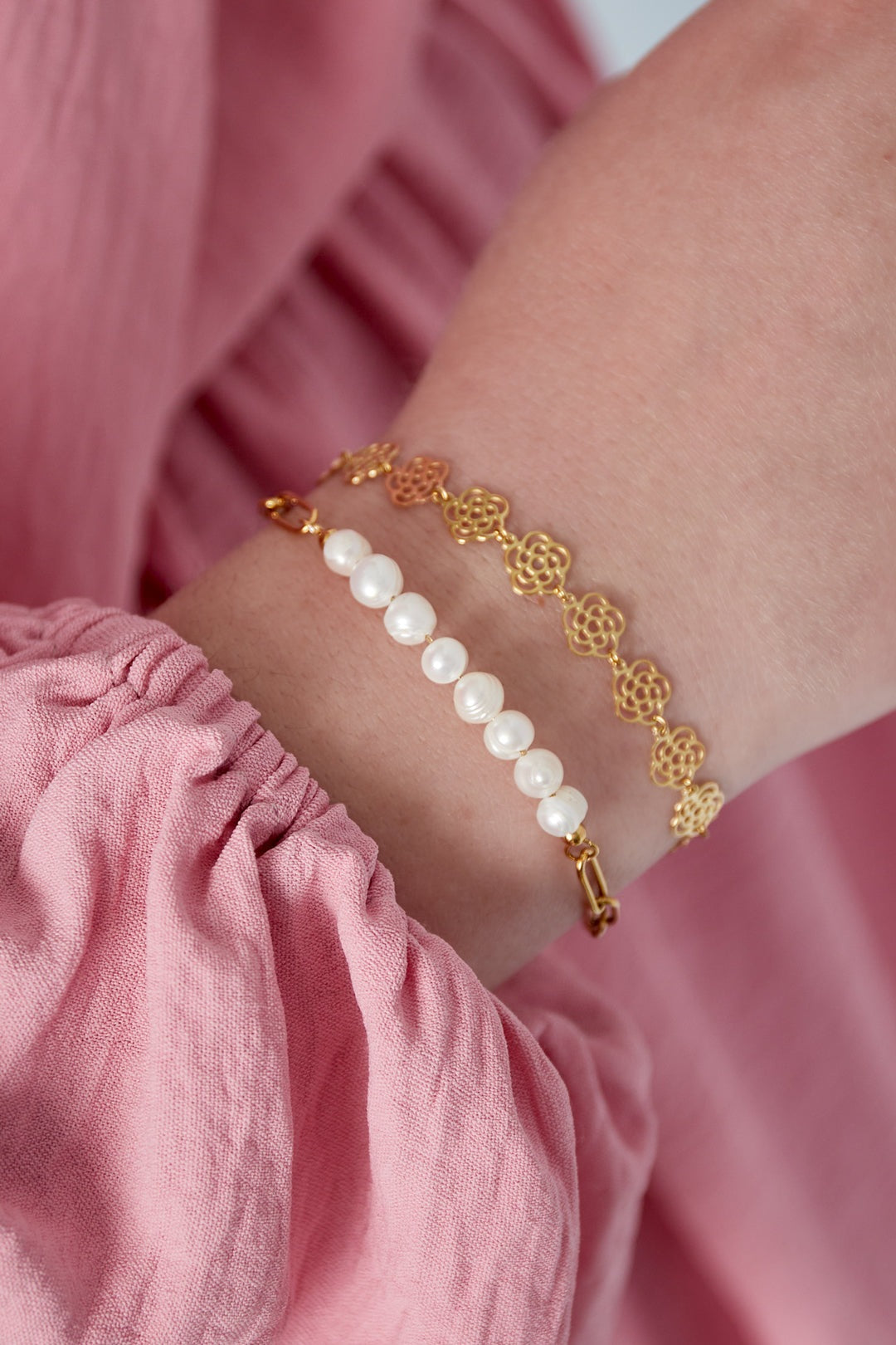 Two bracelets on a woman's wrist - one gold chain bracelet with freshwater pearls and one delicate gold floral chain bracelet, worn with a pink fabric sleeve