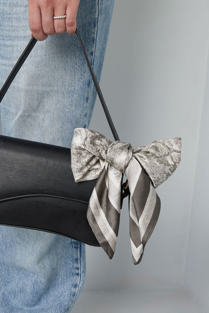 Woman holding a black handbag adorned with a tied scarf bow in gray and white scenic print