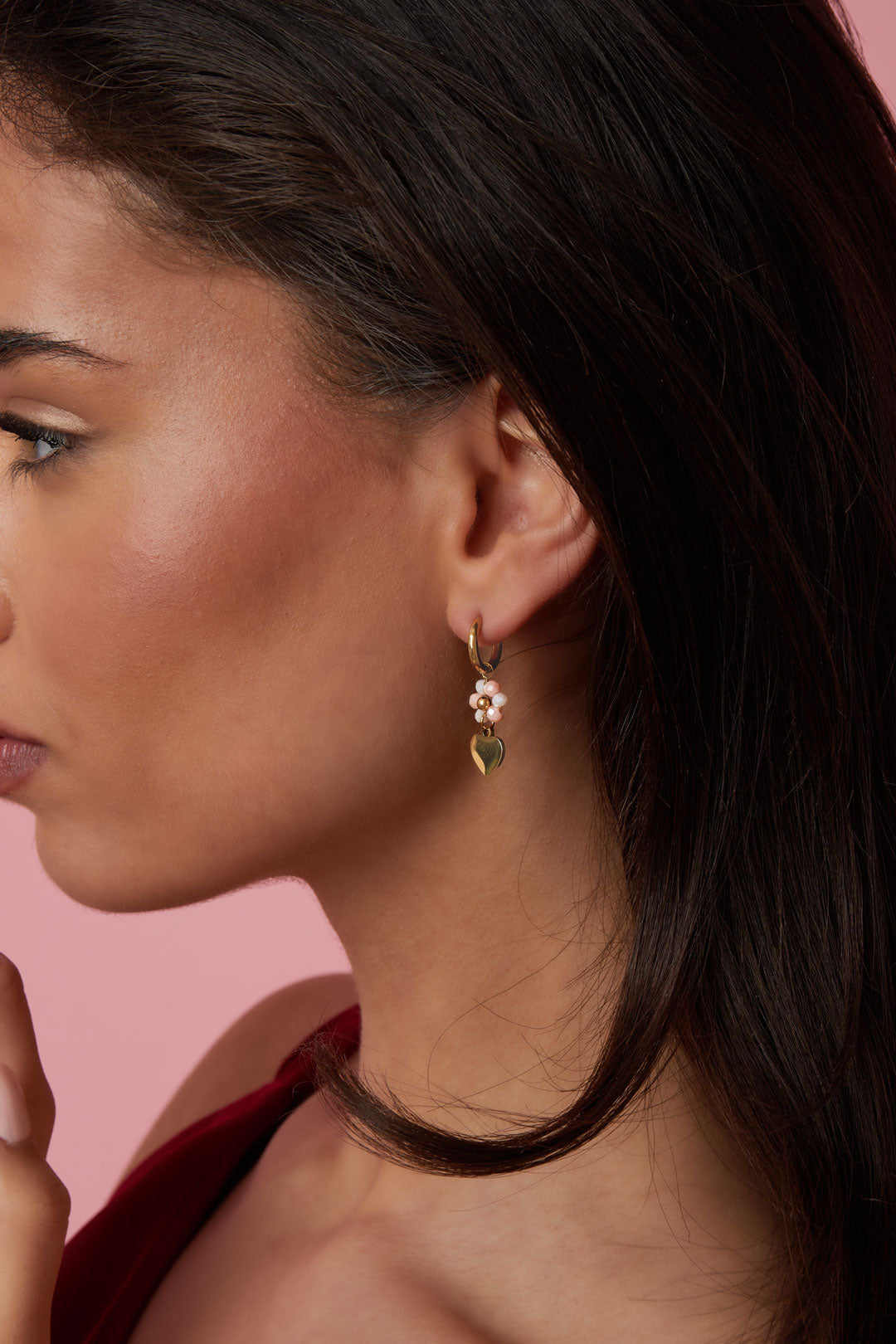 Close-up side view of a woman wearing gold-tone hoop earring with soft pink beads and heart charm against pink backdrop