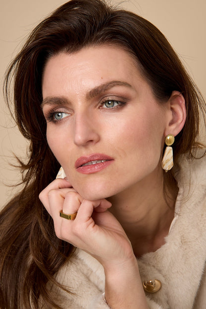 Close-up of woman wearing cream marbled drop earrings with thoughtful expression against beige backdrop