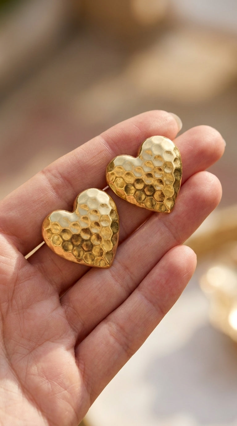 Gold-toned hammered heart stud earrings with a rustic, textured finish held in a hand.