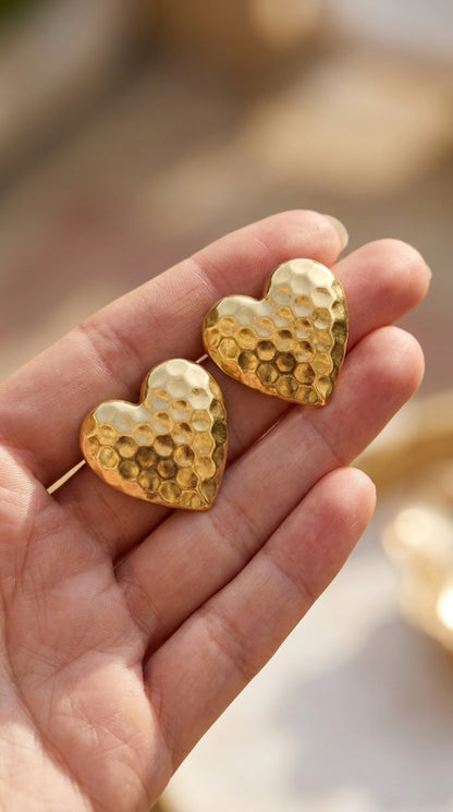Gold-toned hammered heart stud earrings with a rustic, textured finish held in a hand.