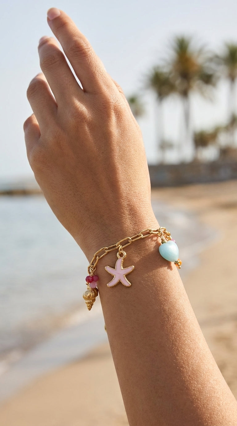 Coastal Charm Stack gold chain bracelet featuring a pink starfish, blue heart, and golden shell charms on a beach.