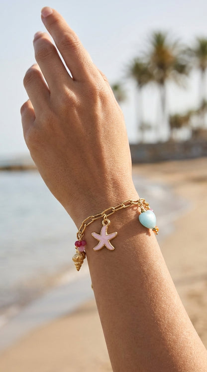 Coastal Charm Stack gold chain bracelet featuring a pink starfish, blue heart, and golden shell charms on a beach.