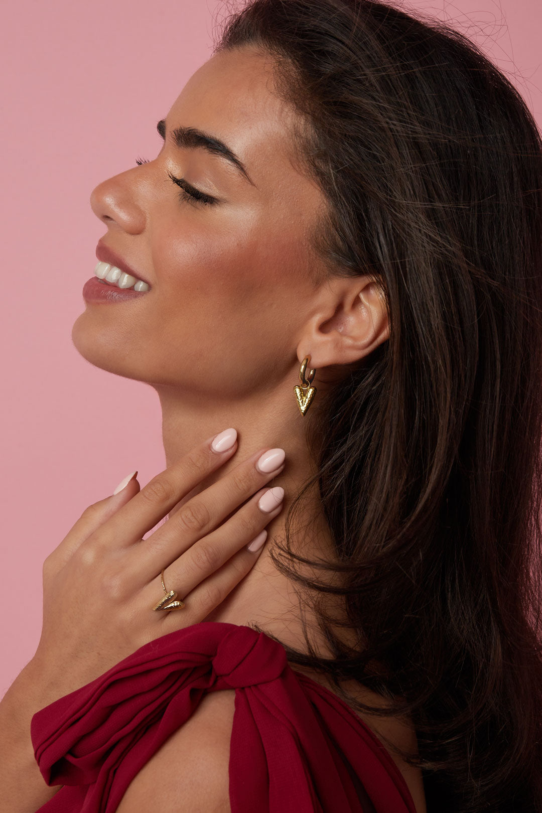 Model wearing gold textured heart drop earrings and matching ring against pink background