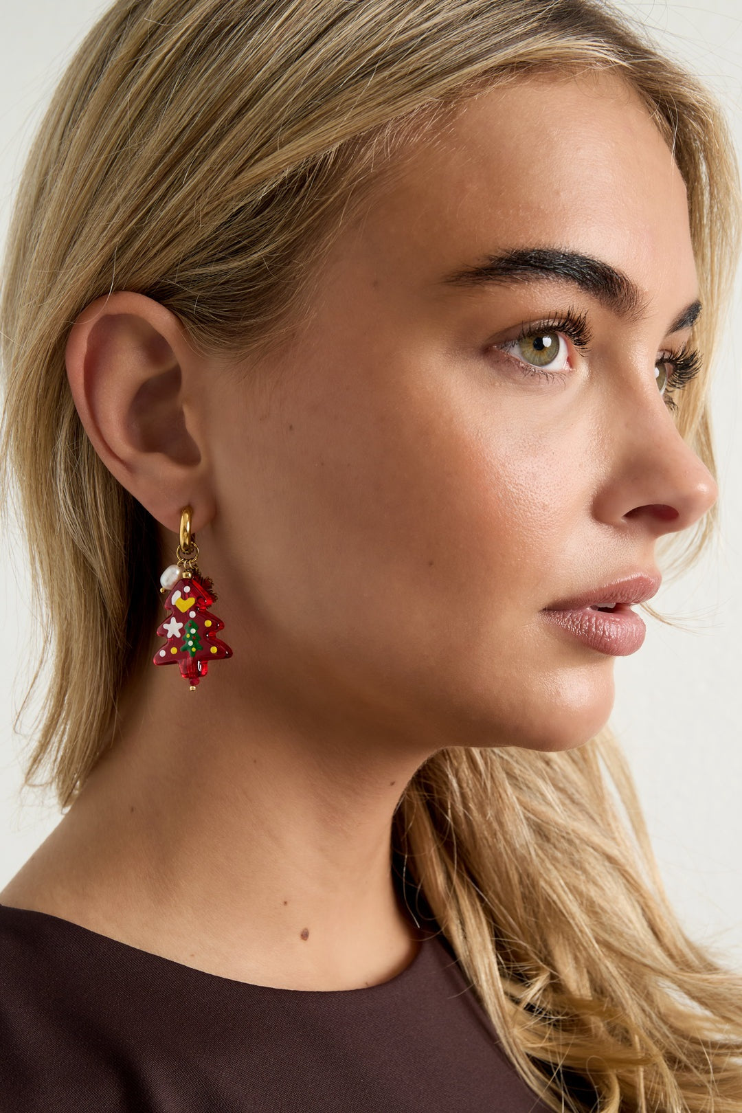 Side profile of a woman wearing the festive Christmas tree drop earring, highlighting the colorful resin charm and gold hoop against her blonde hair and brown top