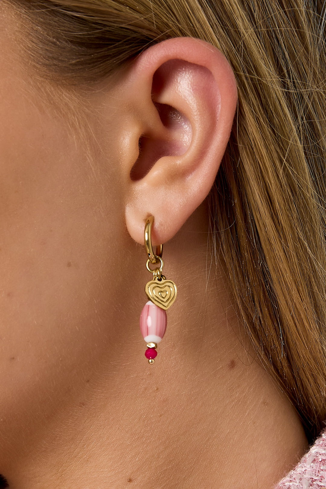 Close-up ear shot showing gold hoop earring with pink beaded charm and gold heart detail on a woman with light skin and blonde hair