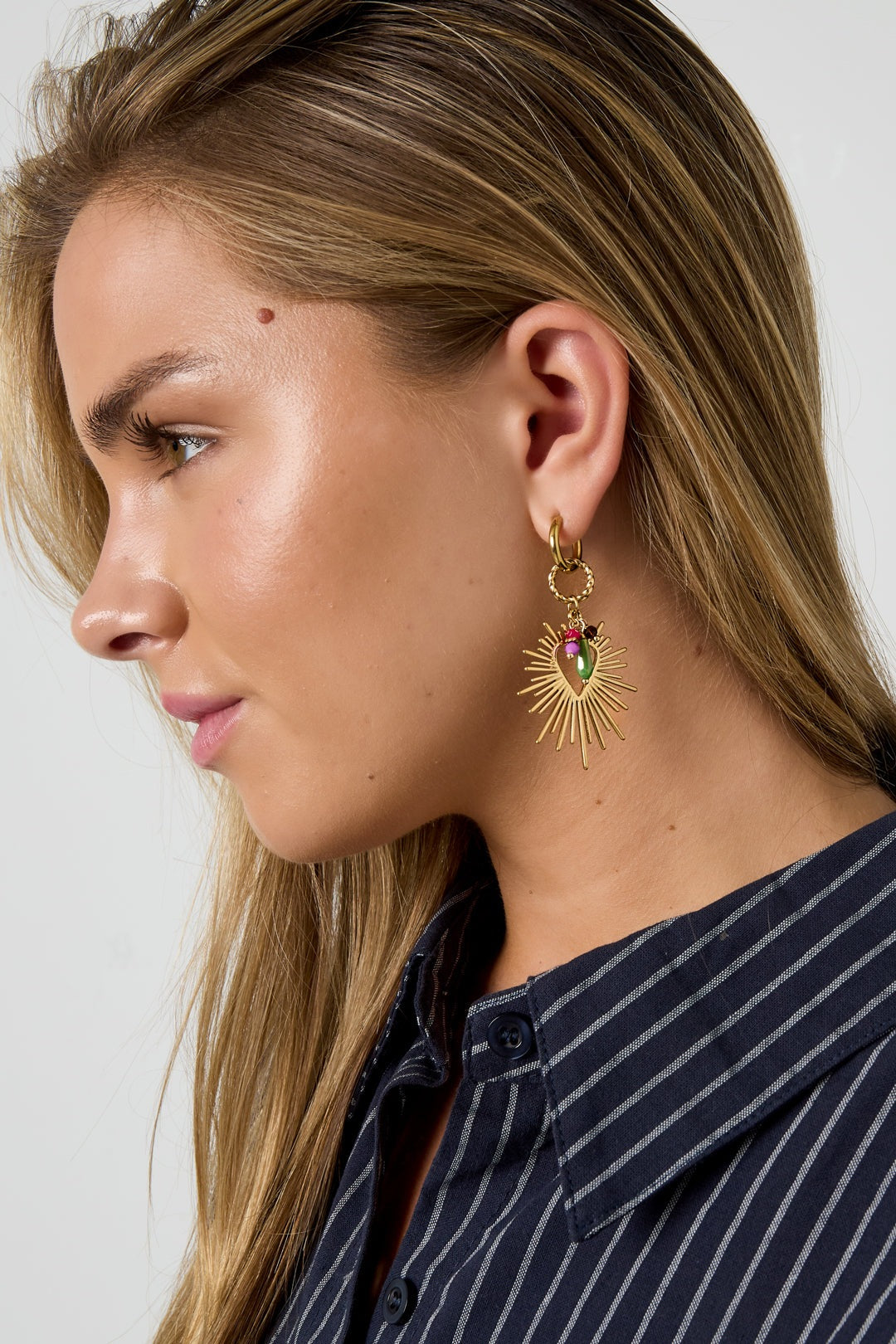 Side profile of woman wearing gold-tone radiant heart charm drop earring showing sunburst and colorful beads
