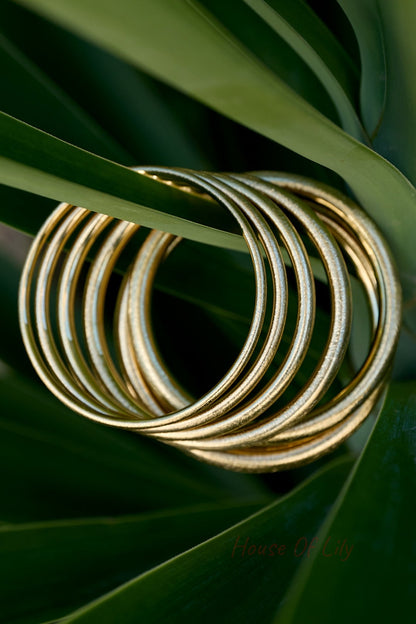 Stack of circular gold bangles with a brushed matte finish, resting on green tropical leaves.
