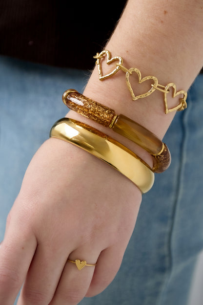 Woman's wrist adorned with three bracelets including a glitter resin bracelet, gold heart link bracelet, and gold cuff, plus a dainty heart ring